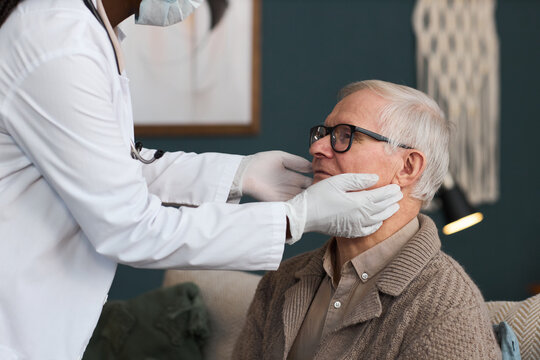 Senior Caucasian man sitting while Black woman doctor wearing gloves examining his jaw and neck during medical checkup in home setting