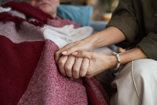 Middle aged Caucasian woman holding hand of senior Caucasian man lying under blanket, showing support and comfort, close up on hands emphasizing caregiving and compassion