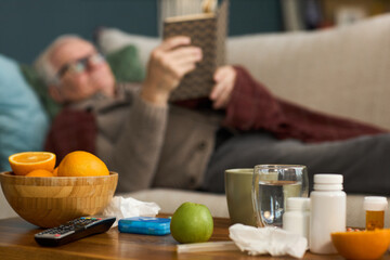 Senior Caucasian man reclining on sofa reading book, foreground showing bowl of oranges, green apple, glass of water, medication bottles, tissues, remote control, mobile phone