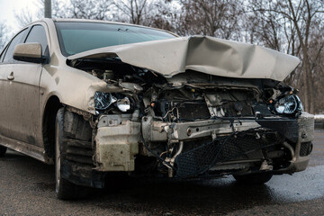 Close-up of the front of a wrecked car after an accident with a badly deformed hood, broken headlights and a damaged radiator on the road.