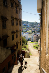 Narrow street in Morocco with tall buildings, a dirt road, and a view of a hillside town. An adult and a child walk uphill in sunlight, with houses and green patches in the distance.
