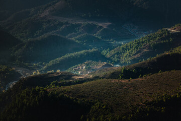Wide view of the Atlas Mountains area in Morocco, with green hills, forested slopes, and sun rays lighting a quiet valley where a small group of houses sits among trees.