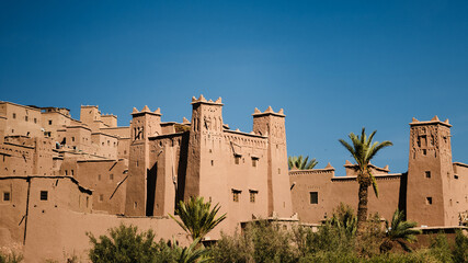 Close view of earthen fortress buildings in Ait Ben Haddou, Morocco, with tall towers, palm trees, and clear blue sky in bright daylight.