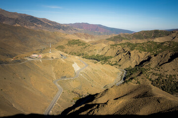 Wide view of the Atlas Mountains area in Morocco, showing dry hills, a winding mountain road, and open valleys under clear blue sky and bright daylight.