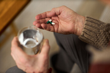 Senior Caucasian man holding glass of water in one hand and several capsules in other hand, preparing to take medication, close up of hands with visible wrinkles and veins