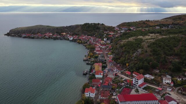 Aerial video of Lin village on Lake Ohrid in Albania, with shoreline houses and soft sun-rays breaking through the clouds.