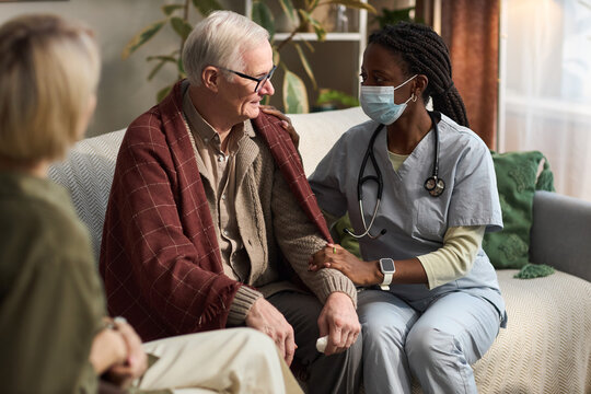 Senior Caucasian man sitting on sofa being comforted by Black female nurse wearing medical scrubs and mask, middle aged Caucasian woman sitting nearby observing interaction