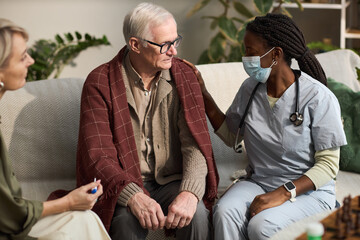 Senior Caucasian man sitting on sofa covered with blanket, receiving support from Black female...