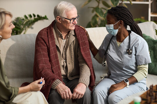 Senior Caucasian man sitting on sofa covered with blanket, receiving support from Black female nurse wearing medical scrubs and mask, while middle aged Caucasian woman observing