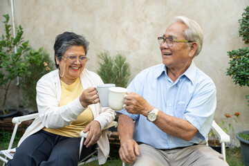 Smiling Asian couple sitting on chair and drinking coffee or tea in garden at home. Happy senior retired relaxing in morning. Healthy old people concept