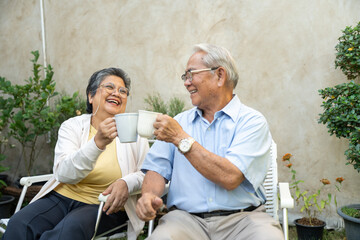 Smiling Asian couple sitting on chair and drinking coffee or tea in garden at home. Happy senior retired relaxing in morning. Healthy old people concept