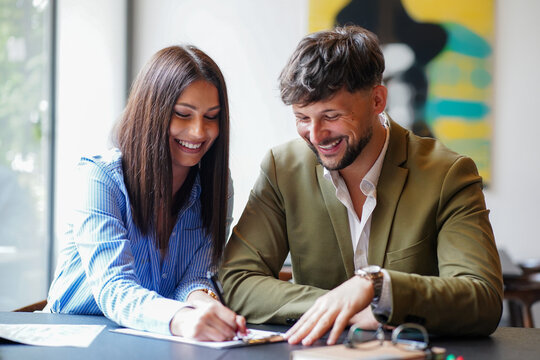 Two people working together at a table in a coffee shop while smiling and sharing ideas during a sunny afternoon