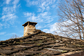 Traditional rural house roof with stone chimney and layered stone tiles under clear blue winter sky