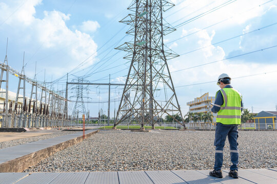 Electrical engineer in safety vest standing at power substation, monitoring high voltage transmission towers and electricity grid infrastructure for maintenance planning and energy security.
