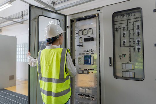 Female electrical engineer in hardhat inspecting indoor switchgear and control cabinet, monitoring power distribution and energy management systems for industrial facility reliability.