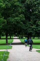 Two people cycling leisurely along a vigeland park path through lush green trees on a sunny summer day. Oslo, Norway