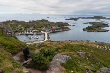 Hiker descending a mountain trail with a full view of a small fishing village, harbor, and numerous islands ballstad lofoten norway