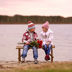 cute children sitting together on a wooden bench on the beach with a bouquet of red roses