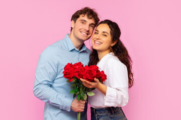 Happy European lady posing with her boyfriend and holding bouquet of red roses, celebrating Valentine's Day together over pink studio background