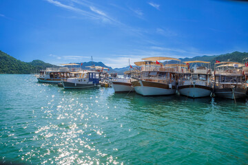 Panoramic wooden pier walkway lined with traditional Turkish gulets and boats flying red flags in a turquoise marina bay. Serene summer scene with green hills and blue skies,