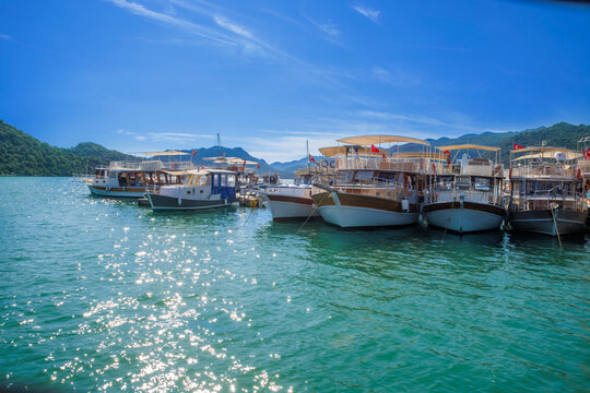 Panoramic wooden pier walkway lined with traditional Turkish gulets and boats flying red flags in a turquoise marina bay. Serene summer scene with green hills and blue skies,