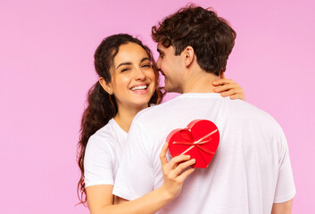 Happy couple in love celebrating Valentine's day, lady holding heart shaped gift box while embracing boyfriend, pink studio background