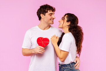 St. Valentines Day. Happy European couple in love embracing, man holding red heart shaped gift box, looking at each other, posing on pink studio background