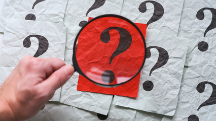 A person with a magnifying glass in his hand magnifying a red piece of paper with a question mark on it. Too many questions. Close up.
