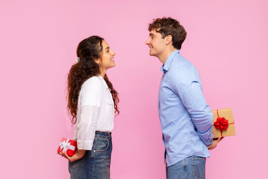 Romantic couple hiding gifts for Valentine's Day or anniversary behind their backs, looking at each other and smiling on pink studio background, side view
