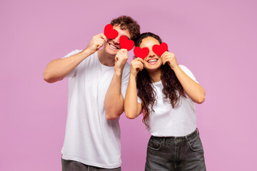 Lovers blinded by their love. Happy couple holding red heart-shaped cards over eyes and smiling, standing over pink background. St. Valentines Day concept