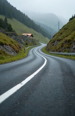 Fototapeta premium Winding road curves through a misty mountain valley towards a small house. Green grassy slopes rise on either side under a cloudy sky. The wet asphalt shows a white dividing line.