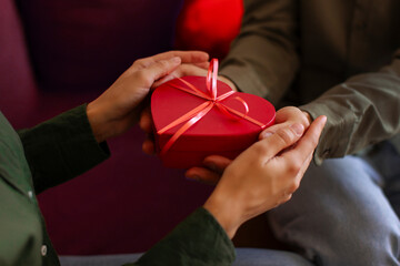 Gift with love. Man giving heart-shaped gift box to his beloved girlfriend, couple sitting on sofa,...