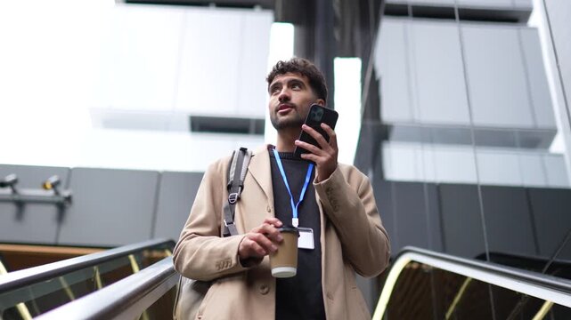 Professional man with an id badge holding a coffee cup and his mobile phone while going up an escalator inside a contemporary corporate office