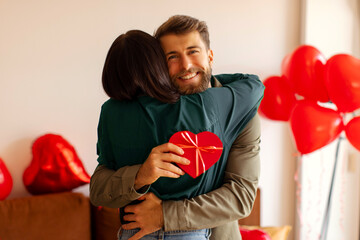 Spouses embracing in warm moment at home surrounded by heart-shaped balloons, exchanging gifts and celebrating Valentine's Day