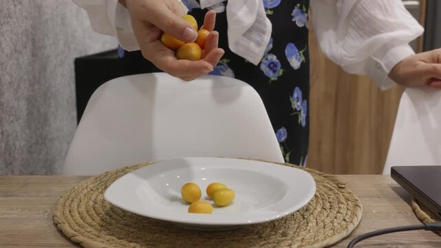 Close up of caucasian woman hands placing kumquat fruits one by one onto white plate on kitchen table, healthy food and minimal domestic scene indoors.