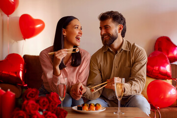 Couple holding chopsticks and sharing sushi, smiling at each other, celebrating anniversary or Valentines day at home with heart-shaped balloons around