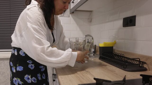 Middle aged caucasian woman in white blouse organizing glassware on wooden countertop near sink and dish rack in modern kitchen, symbolizing housework and tidy lifestyle.