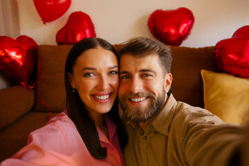 Happy European couple in love taking selfie in decorated with red heart shaped balloons room, celebrating Valentine's day in romantic atmosphere at home