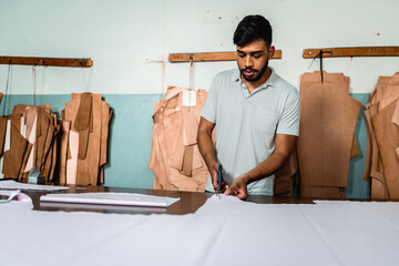 Professional male tailor cutting white fabric with scissors in a garment workshop with clothing patterns in the background.