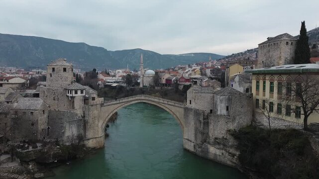 Aerial view of the historic stone bridge over the Neretva River in Mostar, with old town architecture, turquoise water, and surrounding mountains under an overcast sky. Amazing Bosnia and Herzegovina