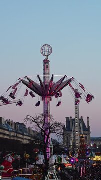 Swing carousel amusement ride operating at the central Christmas market in Paris during winter evening, with festive lights and city architecture in the background.