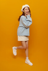 Surprised joyful reaction with Asian woman covering mouth while standing on one leg to express excitement playful emotion fun lifestyle moment on yellow background.