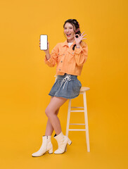 Smartphone app promotion Asian woman holding blank screen mobile phone and showing OK hand gesture while sitting on stool to present digital marketing mobile advertising online engagement.