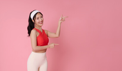Fitness promotion portrait smiling Asian woman in activewear pointing at copy space healthy lifestyle concept workout motivation advertising direction wellness marketing pink studio background.