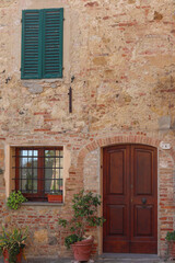 A charming entrance in Montepulciano, Tuscany, featuring a rustic stone wall, a wooden door, and a window with green shutters. Beautiful plants add a touch of warmth to this Italian countryside scene.