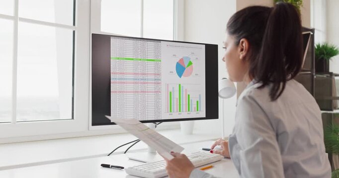 Analyst woman reviews data charts on computer monitor. She reviews an office dashboard and spreadsheet, interprets metrics, and prepares a report for planning. Clear data driven decision concept.