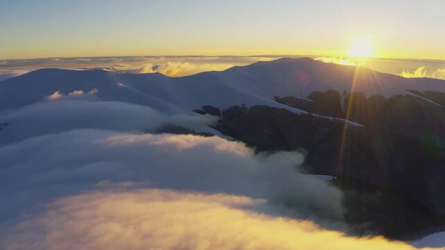 Cloud waves gently and slowly flow over the tops of snow-capped mountains covered with spruce forests at a cold sunset