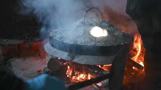 Traditional Albanian Sa&ccedil; Cooking Method with Metal Lid Covered in Hot Ashes Over an Open Wood Fire Hearth in a Rustic Kitchen