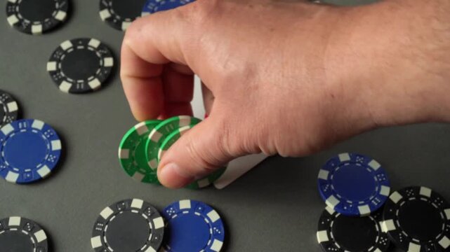 A hand reaches for green poker chips on a tabletop. An ace card is nearby, and black and blue chips are scattered around. It seems to be a gambling scene with mixed colors of chips.