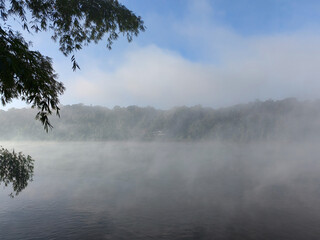 early morning fog rising from calm lake water with lush green trees and hills in background sunlit mist creating serene tranquil atmosphere beautiful natural landscape scenery
